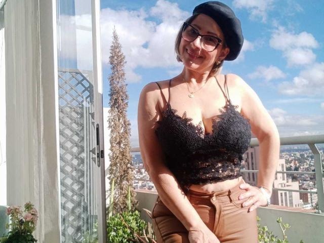 JaneTurner with big boobs, wearing a black lace crop top, brown high-waisted pants, black beret, glasses, and a necklace, standing on a balcony with a city view.