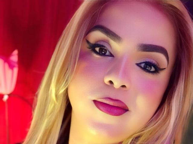 RoxanaLatinaTrans has blonde hair, thick eyebrows, dramatic eyeliner, and pink lipstick. Her face is illuminated with a purple and pink glow. Her ass is not visible.