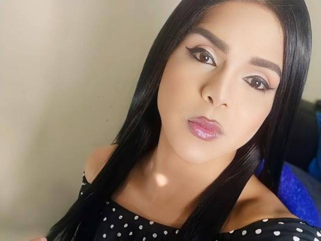 RoxanaLatinaTrans has long black hair, brown eyes, and glossy pink lips. She's wearing a black off-shoulder top with white polka dots. Her makeup includes winged eyeliner.