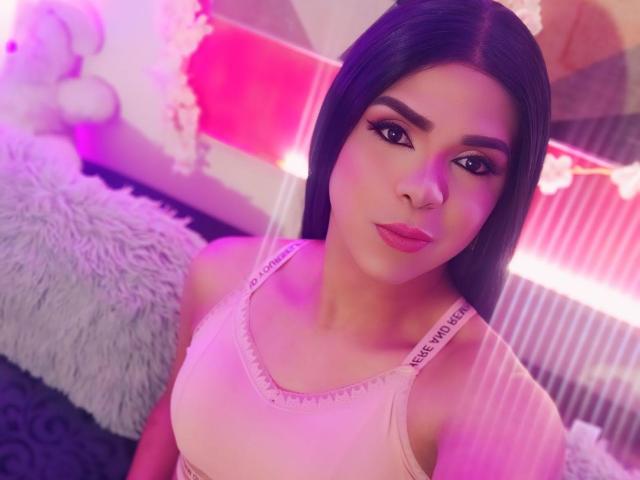 RoxanaLatinaTrans with long black hair, pink tank top, and pink lipstick, looking at the camera with a serious expression. Pink and purple lighting, fluffy white pillow in background.