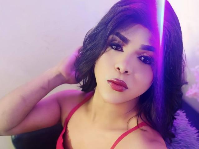 RoxanaLatinaTrans has medium-length black hair, red lipstick, and wears a red top. She's looking at the camera with a slight smile, a purple laser beam on her face.