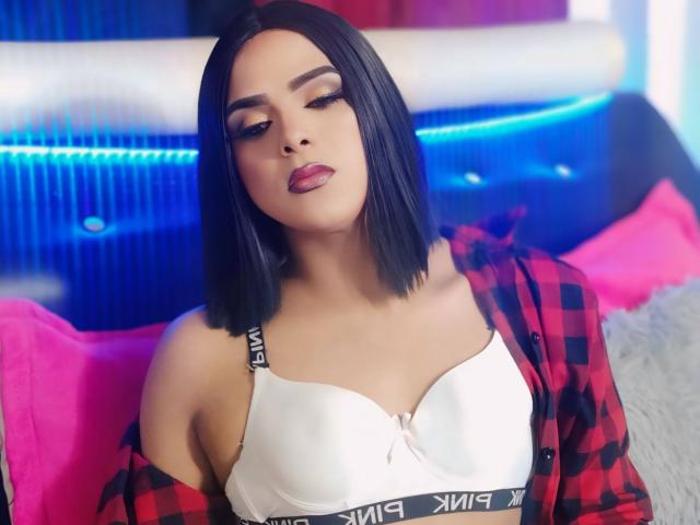 RoxanaLatinaTrans with black bob, wearing white bra, red check shirt, pink pillows, blue neon background, smoky eye makeup, glossy pink lips, and a confident, slightly sultry expression.