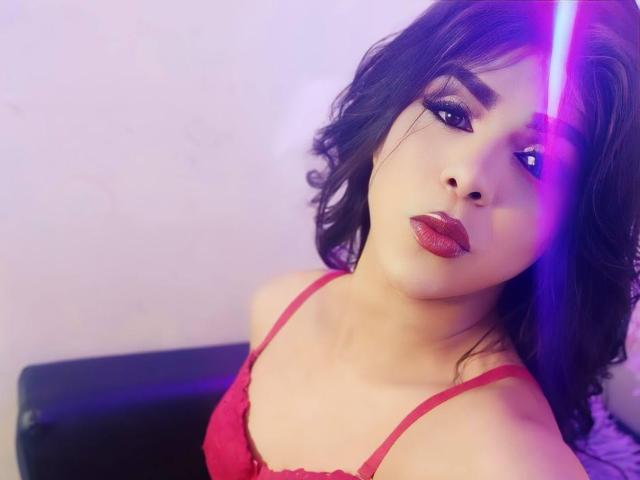 RoxanaLatinaTrans with wavy black hair, red lipstick, and red lingerie, looking up with a purple laser line on her right cheek, showing off her tits and sexy face.