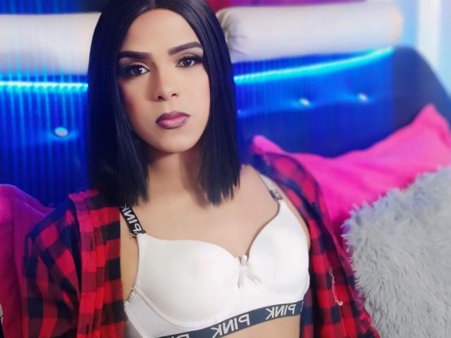 RoxanaLatinaTrans with black bob, wearing red plaid shirt, white bra showing her tits, pink pillow, blue headboard, and gray fluffy pillow, looking directly at camera.
