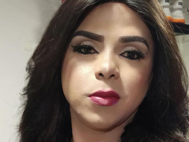RoxanaLatinaTrans has dark brown hair, thick eyebrows, black eyeliner, and pink lipstick. Her face is centered, looking slightly to the right with a neutral expression.