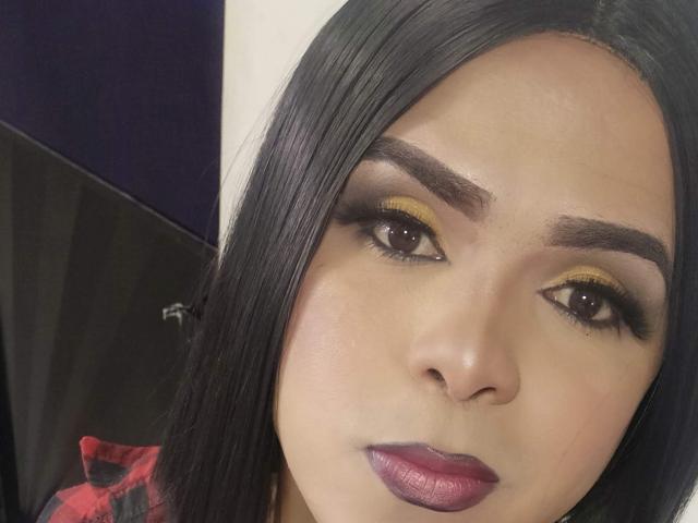 RoxanaLatinaTrans has dark straight hair, yellow eyeshadow, thick black eyebrows, purple lipstick, and a slight smile. Her skin is light brown and she looks directly at the camera.