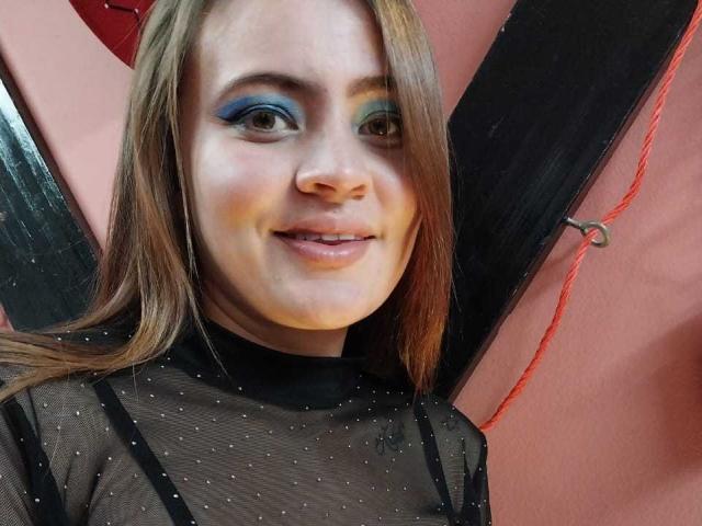 AndrooMedaa with blue eyeshadow, sheer black top, small tits, smiling, brown hair, fair skin, red wall, and black wing-like object in the background.