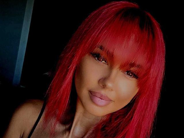 MichelleMarie with bright red, straight hair and bangs, smiling with pink lips, dark eyes, and wearing a black strap top, against a dark background.