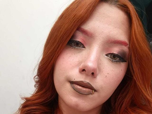 EmmaThomsonn has red hair, freckles, pink and red eyeshadow, brown lipstick with a lip ring, and black eyeliner, looking at the camera with a slight, serious expression.