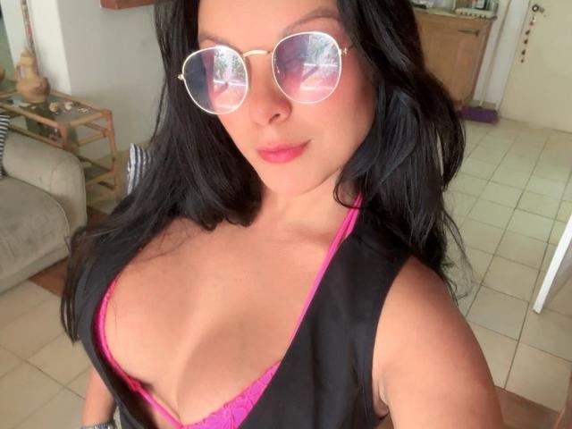 AnyiAnal with big tits, wearing pink lace bra, black vest, and round sunglasses, has wavy black hair, pink lipstick, and a cute smile. Her reflection is in the glasses.