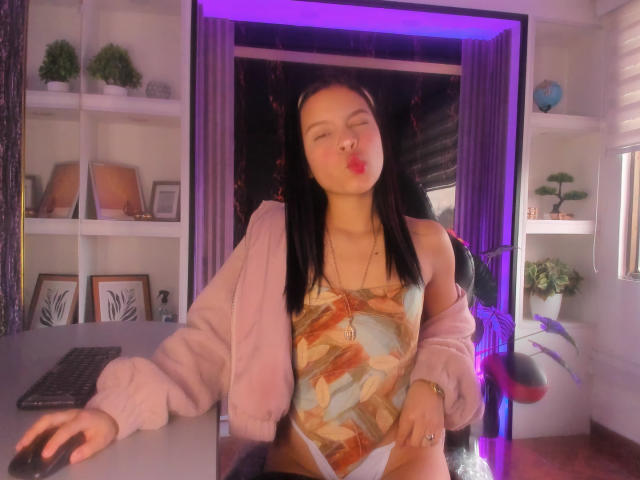 LunaFreya with pouty lips, black hair, pink jacket, and colorful top, leaning on desk, hands on keyboard, purple lights in background, looking cute and sexy.