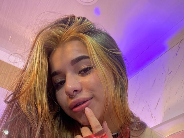 Adhara Evans, com mechas loiras, um piercing no nariz e o dedo tocando os lábios, sorrindo levemente. Sua mão está perto da boca, mostrando seu esmalte rosa. Sua maquiagem é discreta..