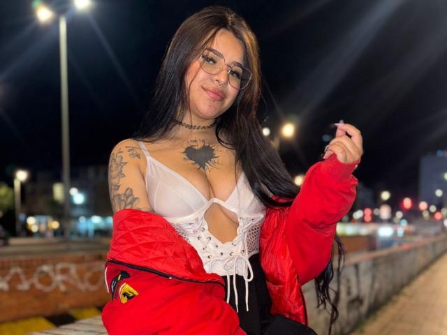 AdharaEvans with long black hair, glasses, red jacket, white lace-up top showing big tits, tattoos, black choker, and black pants, standing at night, holding hair.