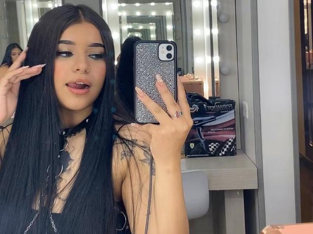 Adhara Evans, com cabelos pretos longos, piercing no nariz e língua de fora, tirando uma selfie em um espelho, segurando um celular com uma capa brilhante, vestindo uma blusa preta, mostrando os seios..