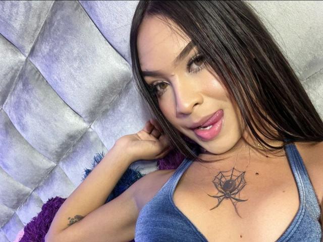 BarbyHadry with long brown hair, blue tank top, tongue out, looking at camera, spider tattoo on chest, small tattoo on arm, lying on gray cushioned surface.