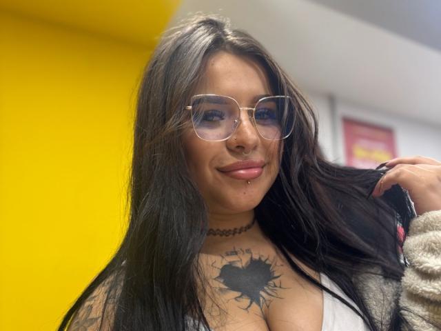 Adhara Evans, com cabelos pretos longos, óculos, piercing no nariz e no lábio, exibindo uma tatuagem em forma de raio solar em seus seios, vestindo um colar e um casaco de pele, sorrindo..