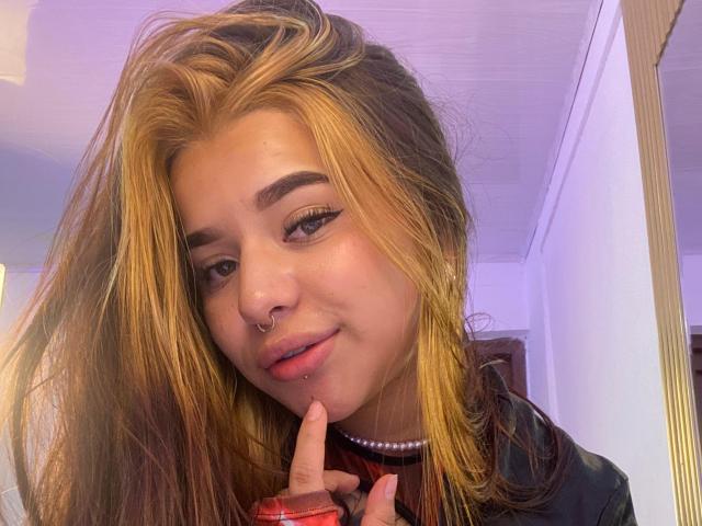 Adhara Evans, avec ses cheveux blonds et bruns, son anneau au nez et son collier de perles, touchant ses lèvres avec son doigt, vêtue de rouge et de noir, a un visage mignon, séduisant et attrayant..