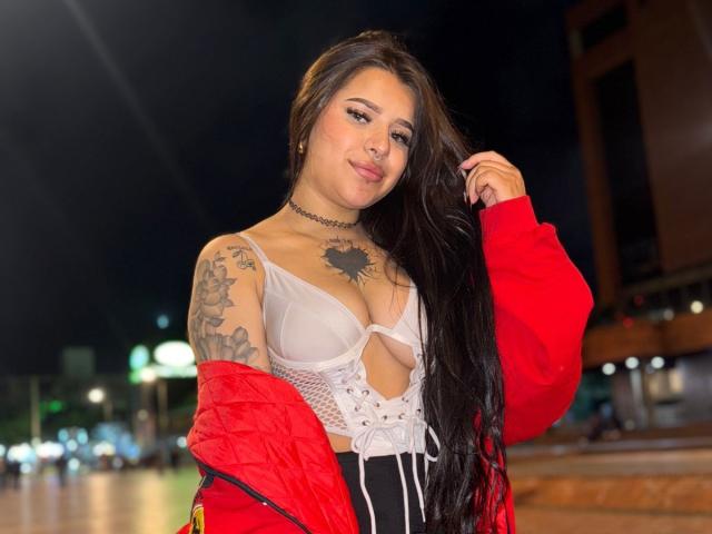 Adhara Evans met lang zwart haar, een rode jas, een witte kantbeha die haar borsten laat zien, tatoeages op haar arm en borst, een zwarte choker, 's nachts buiten, haar vasthoudend, een lichte glimlach..
