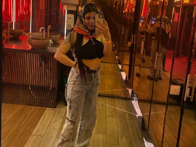 Adhara Evans, met lang donker haar, draagt een zwarte crop top, een hoofddoek en een broek met hoge taille, en maakt een spiegelzelfportret, waarbij haar borsten en billen zichtbaar zijn..