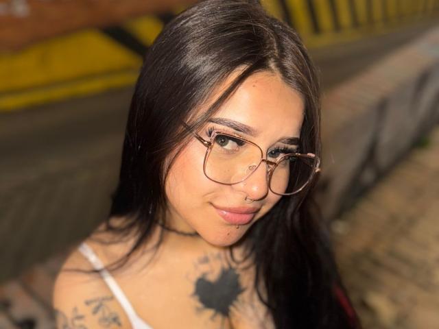 AdharaEvans has long black hair, glasses, a nose ring, tattoos, and a white tank top, showing a cute, slightly smug face with pink lips and brown eyes.