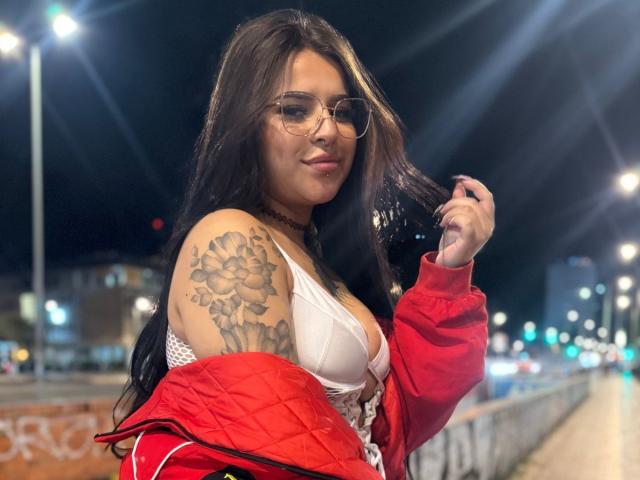 AdharaEvans with long dark hair, glasses, red jacket, white bra showing her big tits, and a tattooed arm, holding her hair, night, street lights, slight smile.