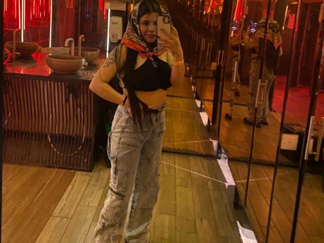AdharaEvans met een gemiddelde borstmaat, draagt een zwarte crop top, een hoofddoek met een patroon en high-waist jeans met slijtage, en maakt een spiegelzelfportret in een donkere kamer met rood licht..