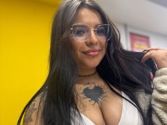 Adhara Evans, con cabello negro largo, gafas, escote blanco, tatuaje de corazón negro en el pecho, collar, piercing en el labio, sosteniendo el cabello, sonriendo, fondo amarillo. Pechos, tatuaje, aro en el labio, linda..