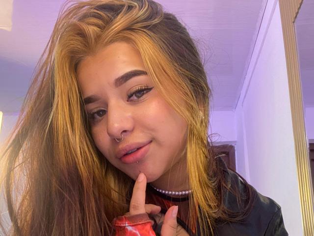 Adhara Evans, con cabello rubio y castaño, aro en la nariz y collar de perlas, labios ligeramente entreabiertos, dedo cerca del mentón, camisa negra, delineador de ojos sutil, sonrisa suave, en interiores, iluminación suave..