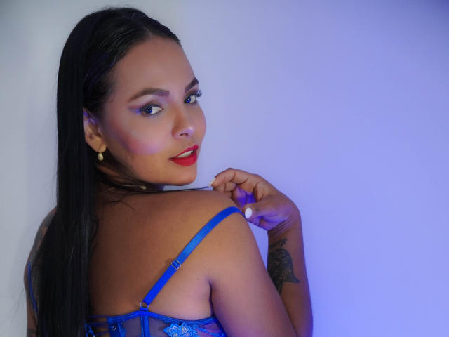 JessicaFiore with long black hair, blue lace bra, red lipstick, tattoo on right arm, looking over shoulder, hand near shoulder strap, blue lighting, smiling. Boobs, tits, cute.