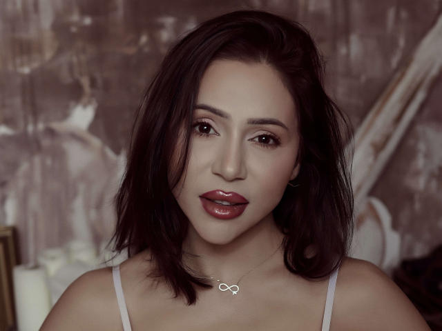 GinaMence has dark brown hair, red lipstick, and a white tank top. She's wearing a silver infinity necklace, with a soft, seductive look. Boobs, ass, pussy not visible.