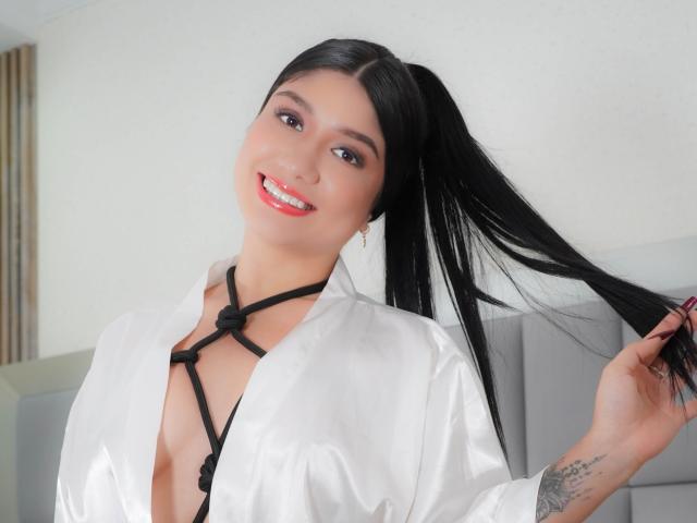 Camila Verone, con cabello negro recogido en una coleta, sonriendo, vestida con una camisa blanca brillante con detalles negros tipo tiras, sosteniendo su cabello, mostrando un tatuaje en su muñeca derecha..