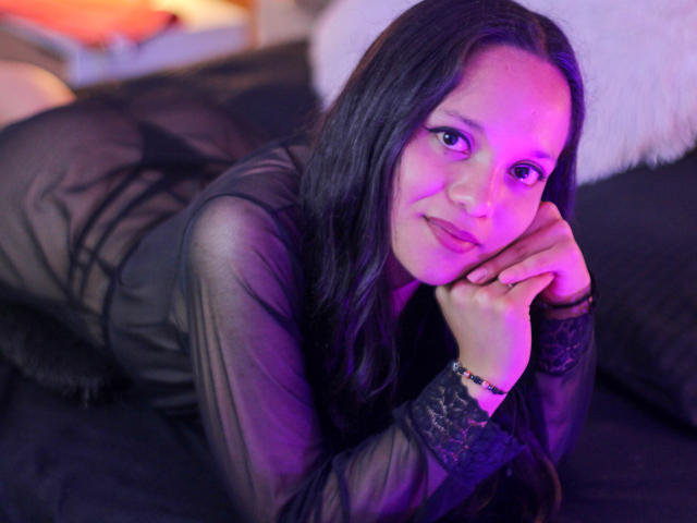 NanamiBrown with long black hair, wearing a sheer black dress, lying on a black bed, hands under chin, purple light, smiling, looking at camera, purple highlights on face.