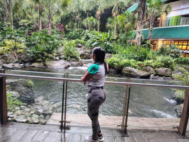 PauletteLoveX, with black hair in a ponytail, green and white top, and grey floral pants, stands on a wooden bridge, looking at a flowing stream, surrounded by lush greenery.