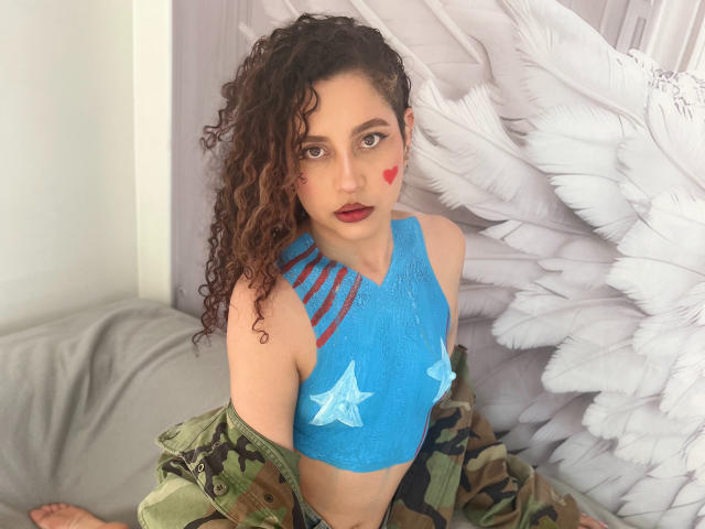 IrinaSweett with curly brown hair, blue star-patterned top, red heart on cheek, camo jacket, and feather wall. She's looking at the camera with red lipstick.