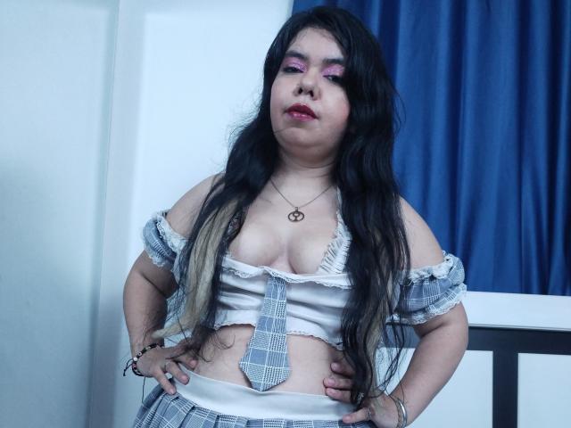 KarlaSexyHott with long black hair, pink eyeshadow, and a plaid crop top showing her boobs, hands on hips, blue curtains in background, wearing a tie and necklace.