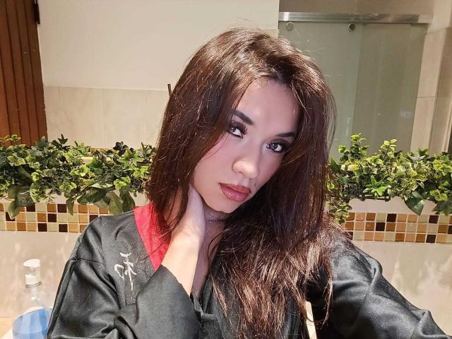 LaylaRousee with long brown hair, wearing a black silk robe with red trim, touching her cheek, looking at the camera with dark eyes, slightly parted lips.