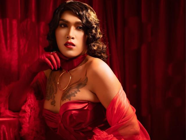 TemptressBella69 with red curly hair, red lipstick, red dress, red gloves, and a tattooed chest, holding a gold ring, looking seductively at the camera.