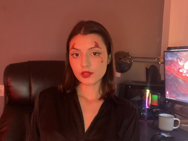 SunnyLeila with dark brown hair, red makeup on forehead, black shirt, red lips, sitting in front of a computer, cup on desk, dim red lighting.