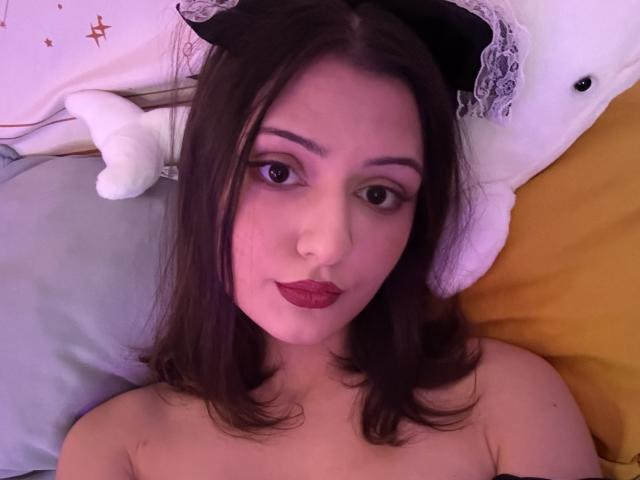 SunnyLeila with dark brown hair, wearing red lipstick, black headband, lying on bed, looking at camera, topless, soft purple and yellow light, white stuffed animal on pillow.