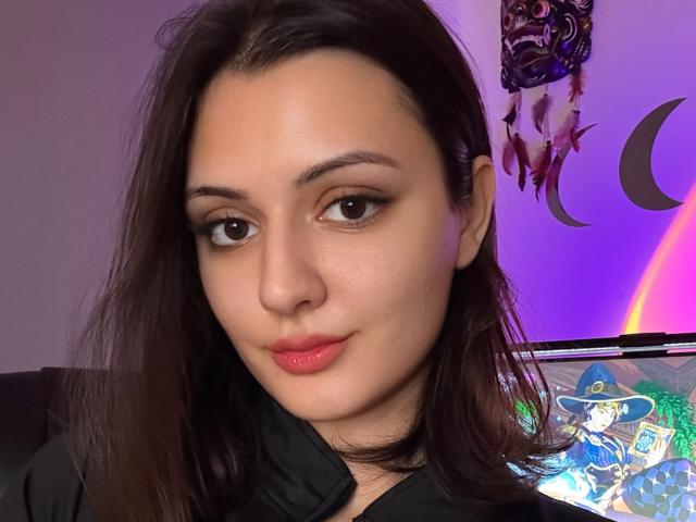 SunnyLeila with dark brown hair, red lipstick, and black shirt, looking at camera with subtle smile. Her face is slightly flushed. Her ass is not visible.