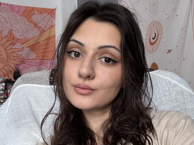 SunnyLeila has wavy dark brown hair, brown eyes with black eyeliner, and pink lips. She's wearing a white top, looking straight at the camera with a neutral expression.