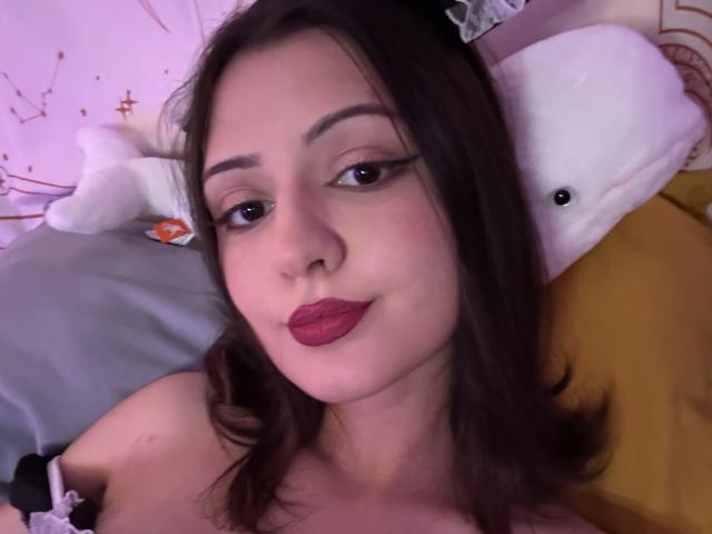 SunnyLeila has dark brown hair, red lipstick, and black eyeliner. She's lying on a bed with a white stuffed whale and yellow pillow. Boobs partially visible.