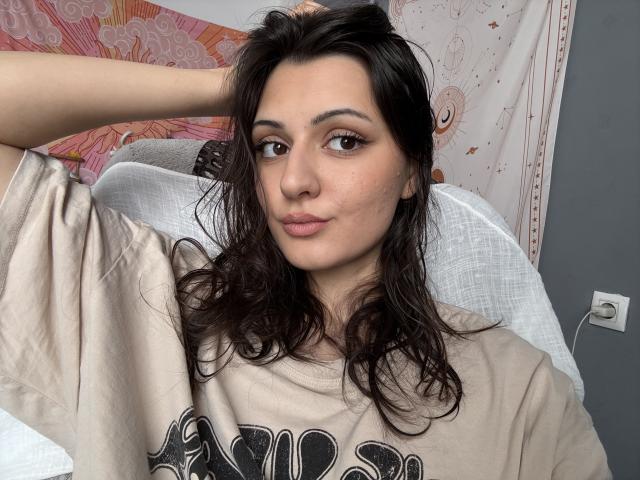 SunnyLeila with wavy dark brown hair, wearing a beige shirt, has her arm raised, looking at the camera with a slight smile, her brown eyes are half-open.