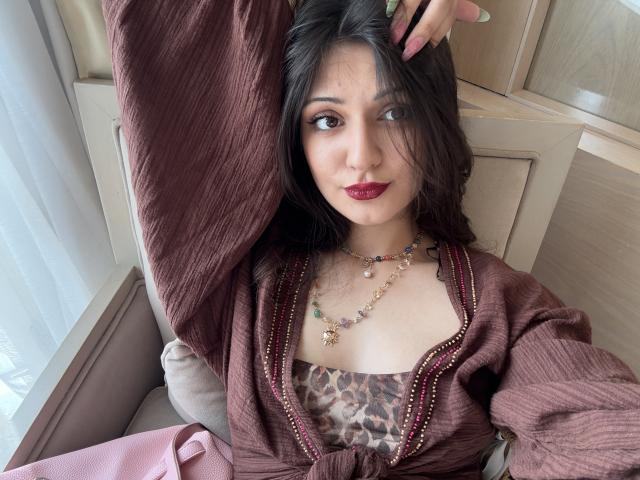 SunnyLeila with dark hair, red lipstick, and a cute face, wearing a maroon cardigan and leopard print top, holding her hair up, showing her necklaces. Boobs slightly visible.