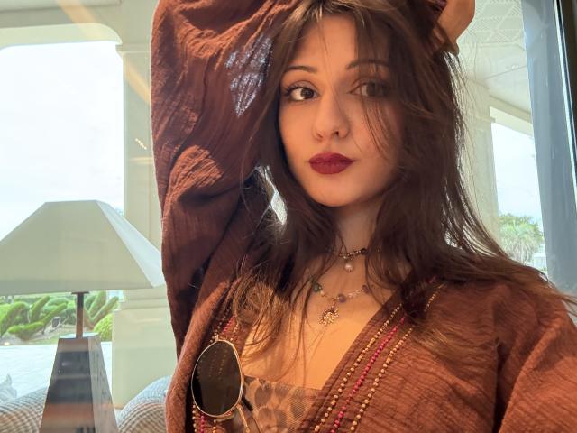 SunnyLeila with brown hair, red lipstick, and a brown cardigan, wearing leopard print and bead necklaces, raising her arm, sunlight in her face, holding sunglasses.