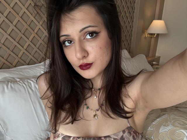 SunnyLeila with dark brown hair, red lipstick, and necklace, taking a selfie, showing her tits in a patterned top, with a slightly flushed face and arm raised.