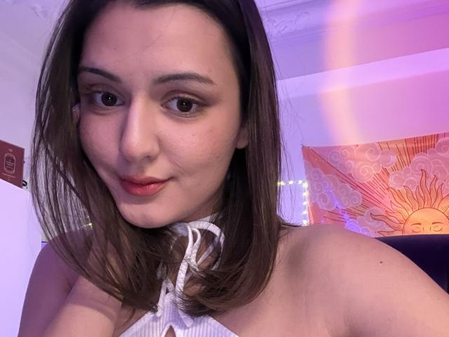 SunnyLeila with shoulder-length brown hair, white halter top, and light skin, smiling at the camera, showing her cute face and subtle makeup, with a hint of pink lighting.