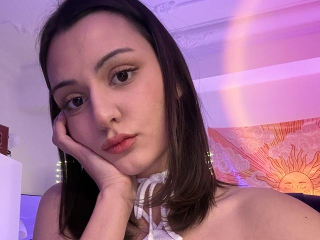 SunnyLeila with brown hair, pouty lips, and fair skin, resting her cheek on her hand, wearing a white halter top, in a softly lit room with purple and pink hues.