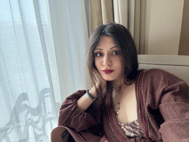 SunnyLeila with brown hair, red lipstick, and a brown cardigan, wearing a necklace and bracelet, sitting by a window, looking at the camera. Her tits are visible.
