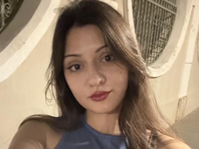 SunnyLeila with long brown hair, red lips, and a blue halter top, showing a calm, slightly pouty face, in a soft, warm-lighted selfie.